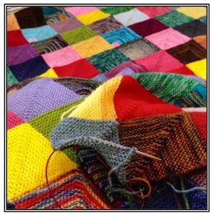 Op de afbeelding: Een kleurrijk gebreid deken met verschillende vierkanten in verschillende kleuren, waaronder rood, geel, blauw, groen en paars. Het deken wordt bewerkt met een breinaald en garen.