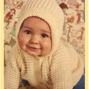 May include: A baby wearing a white knitted hooded sweater and matching trousers. The sweater has a textured pattern and the hood is pulled up over the baby's head.