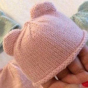 Könnte beinhalten: Eine rosa gestrickte Baby-Mütze mit Bärenohren. Die Mütze ist aus weichem, strukturiertem Garn gefertigt und hat ein schlichtes, klassisches Design.