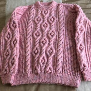 Peut inclure: Un pull rose tricoté à la main avec un col rond. Le pull présente un motif en tricot torsadé avec des motifs de losanges et de cordes. Les manches ont également un motif en tricot torsadé. Le fil a des mouchetures de rose et de violet plus foncés.