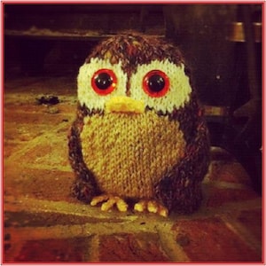 May include: Hand-knitted owl figurine with large red eyes, a yellow beak, and a textured, multi-colored body. The owl is made of yarn and sits on a brick surface. A charming, handmade decorative item.