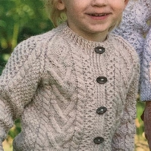 May include: A light beige knitted cardigan with a cable knit pattern and button closure. The cardigan is being worn by a young child.