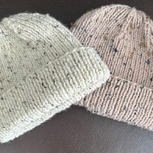 May include: Two knitted hats, one white with flecks of brown and one pink with flecks of brown and blue.
