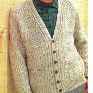 May include: A beige V-neck cardigan featuring a textured knit pattern, long sleeves, ribbed cuffs, and a front button closure with six brown buttons. The garment includes two front pockets and is worn over a dark green plaid button-down shirt.