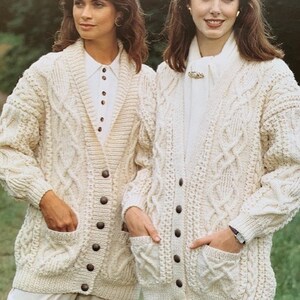 May include: Two women wearing cream-colored cable knit cardigans with button closures and pockets. The cardigans have a shawl collar and are worn over white shirts.