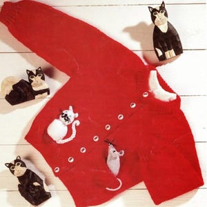 May include: A bright red cardigan sweater with white buttons and cat and mouse appliqués. The sweater is surrounded by three wooden cat figurines in black and white. The sweater features a white cat and a gray mouse.