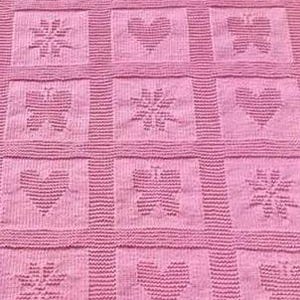 Op de afbeelding: Een roze gebreide deken met een herhalend patroon van vierkanten. Elk vierkant heeft een hart-, vlinder- of sneeuwvlokontwerp. De deken heeft een getextureerd uiterlijk en een zachte, pastelkleur.