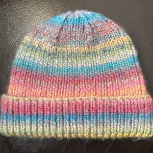 May include: A colorful striped knitted beanie hat with a folded brim. The hat is made with a variety of shades of blue, pink, and yellow yarn.