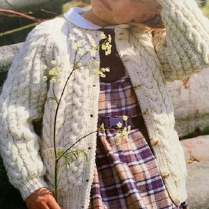 May include: A white cable knit cardigan with a button closure and a patch pocket. The cardigan is worn over a plaid dress.