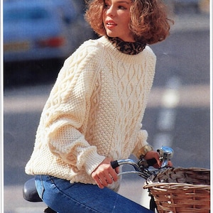 May include: A woman wearing a white cable knit jumper and blue jeans is riding a bicycle with a wicker basket.