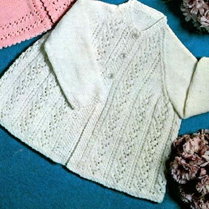 May include: Cream-coloured, knitted baby cardigan with a delicate openwork pattern. The cardigan features long sleeves, a rounded collar, and three small buttons. A pink knitted piece is visible in the background.