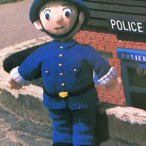 May include: A knitted doll dressed as a police officer, wearing a blue uniform with gold buttons and a yellow star on the helmet. The doll has a smiling face and is standing in front of a brick building with the word "POLICE" on it.