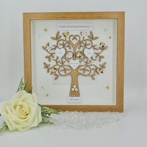 May include: A wooden frame with a white background and a gold tree with white flowers and hearts. The tree has the names "Tim" and "Lisa" on it, as well as the date "26th February 1967". The frame also has the text "Golden Wedding Anniversary" and "50 Years".