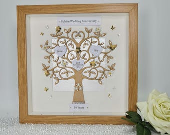 May include: A square wooden frame containing a decorative tree cutout with gold and white accents. The tree features three heart-shaped labels with the text James, Ellie, and 21st November 1974. A banner at the top reads Golden Wedding Anniversary.