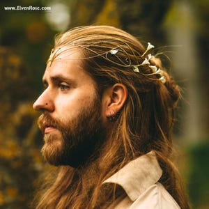May include: A gold-toned headpiece with delicate floral and leaf accents. The headpiece is worn on the head of a person with long, light brown hair and a beard. The headpiece is a decorative accessory.