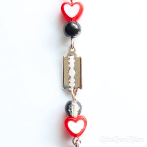 May include: A close-up of a silver razor blade charm with red and white heart beads and black beads on a chain.