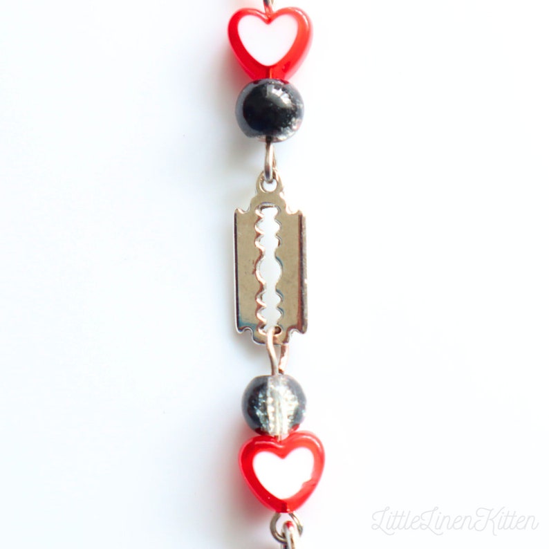 May include: A close-up of a silver razor blade charm with red and white heart beads and black beads on a chain.