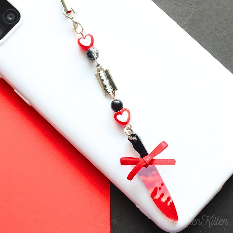 May include: A red and black phone charm with a knife, hearts, and a razor blade. The knife has a red and white design that looks like blood.