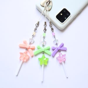 May include: Three pastel phone charms with star-shaped lollipops. Each charm features a bow, a star-shaped lollipop, and a beaded chain. The colours include pink, green, and purple. The charms are attached to a white phone.