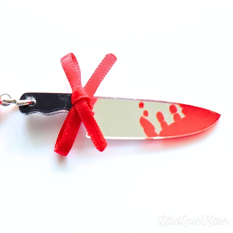 May include: A red and black knife keychain with a red bow and a blood splatter design. The keychain is made of acrylic and has a silver chain.
