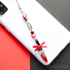 May include: A red and black knife charm with a red bow and a red heart charm with a black bead. The charm is attached to a white phone case with a black and white phone.