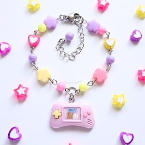 May include: A pastel bracelet with a pink game controller charm. The bracelet features pink, yellow, and lilac beads in flower, heart, and round shapes. The game controller charm has a "Win" screen with bear and bunny graphics.