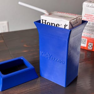 Op de afbeelding: Blauwe plastic houder voor een sapdoos met een afneembare basis. De houder heeft de tekst "KidKuzee" erop gedrukt. Een sapdoos zit in de houder met een rietje dat eruit steekt.