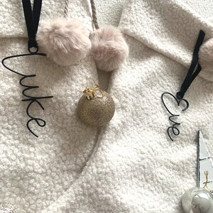 Può includere: Primo piano di due calze di Natale bianche strutturate con targhette nere con i nomi "Luke" ed "Eve". Ogni calza ha un soffice pompon rosa e un ornamento dorato glitterato. È visibile anche un ornamento argentato.