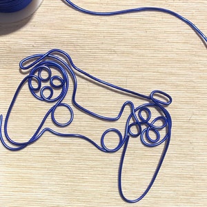 May include: A blue wire sculpture depicting a games controller. The controller is crafted from a single length of blue wire, with details such as buttons and joysticks. A spool of blue wire is visible in the upper left corner.