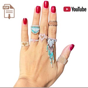 Może przedstawiać: Dłoń prezentuje kilka pierścionków z koralików w różnych wzorach. Pierścionki mają różne kolory, w tym złoty, turkusowy, różowy i biały. Niektóre pierścionki mają kształt serca, inne frędzle. Paznokcie są pomalowane na czerwono.