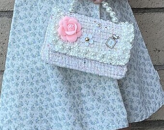 May include: A pink and white tweed purse with a pearl handle and a pink flower embellishment. The purse has a small gold charm with the letter "S" on it.