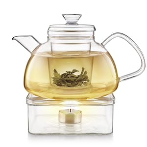 May include: A clear glass teapot with a built-in infuser, filled with tea and tea leaves. The teapot sits on a clear glass warmer with a lit candle inside. The teapot has a curved handle and a spout.