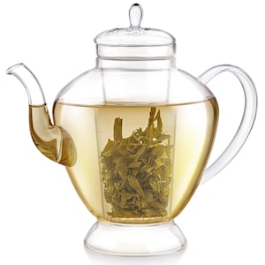 May include: A clear glass teapot with a curved spout and handle. The teapot is filled with a light yellow liquid and green tea leaves. The lid has a small handle. The teapot sits on a clear base.