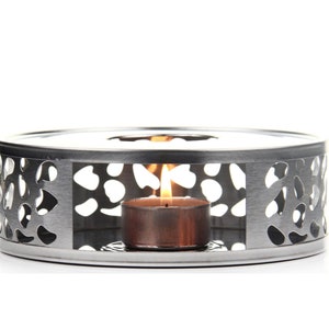 May include: Silver tea warmer with a decorative cutout design and a lit tea light candle in the center.