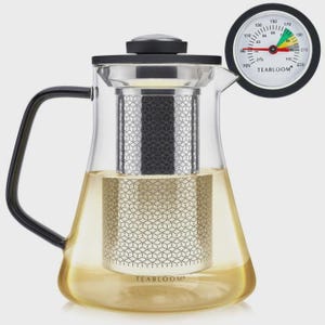 May include: A clear glass teapot with a black handle and lid, featuring a stainless steel infuser with a geometric pattern. The word TEABLOOM is printed on the glass. A round analog thermometer with a white face and black rim is positioned nearby.