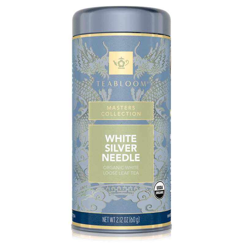 Op de afbeelding: Cilindrische theebus met een lichtblauwe achtergrond en gouden accenten. Het etiket luidt "TEABLOOM MASTERS COLLECTION WHITE SILVER NEEDLE ORGANIC WHITE LOOSE LEAF TEA." Nettogewicht 60 g.