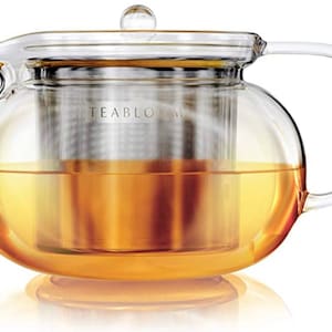 May include: A clear glass teapot with a stainless steel infuser. The teapot is filled with a golden-colored tea. The teapot has a glass lid and a glass handle. The text "TEABLOOM" is printed on the side of the teapot.