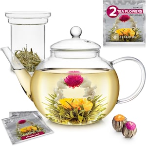 May include: A clear glass teapot with a flowering tea bloom inside. The teapot is round with a handle and a lid. A glass infuser with loose tea is to the left. Two tea flower sachets and two tea flowers are also included.