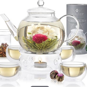May include: A clear glass teapot with a flowering tea bloom inside, surrounded by four double-walled glass teacups. A small candle warmer sits beneath the teapot. A container of loose tea and a box of flowering teas are also present.