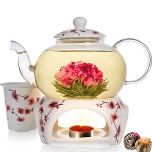 May include: A white ceramic teapot with a glass body and a floral design. The teapot is filled with a blooming tea flower and sits on a white ceramic warmer with a candle.