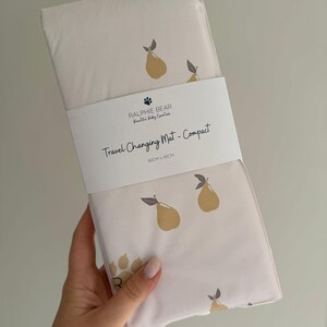 May include: A cream-colored travel changing mat with a pear pattern. The mat is folded and wrapped with a white band that reads "RALPHIE BEAR Beautiful Baby Essentials" and "Travel Changing Mat - Compact". The mat measures 60cm x 40cm.