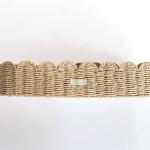 May include: A round woven basket with a scalloped edge. The basket is made of natural-coloured fibres and has a small, rectangular metal tag in the centre. Ideal for storage or display, it is a decorative item.