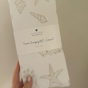 May include: A compact travel changing mat featuring a white backdrop adorned with light brown seashells and starfish. The mat is 60cm x 40cm. The packaging displays the text "RALPHIE BEAR Beautiful Baby Essentials" and "Travel Changing Mat - Compact".