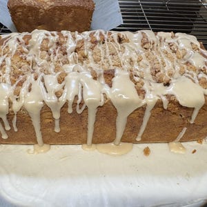 May include: A rectangular loaf of golden-brown baked bread topped with a crumbly streusel texture and drizzled with a thick, opaque beige glaze. The loaf sits on a piece of parchment paper, with another smaller loaf visible in the background.