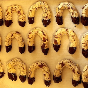 May include: A baking sheet filled with almond crescent cookies dipped in chocolate. The cookies are arranged in rows and are all the same size and shape.