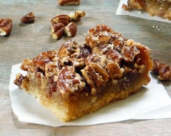 May include: A square slice of pecan pie on a white paper square. The pie has a golden-brown crust and a topping of caramelized pecans. Scattered pecans are on the wooden surface around the pie.