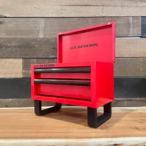 May include: A bright red metal tool chest with two drawers and a hinged lid. The text US GENERAL is printed in black on the front of the lid and the drawer frame. The unit is supported by four black U-shaped legs, providing a stable base for storage.