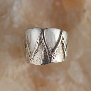 May include: A silver ring with a textured, geometric design. The ring is made of metal and has a brushed finish.
