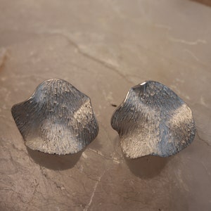 May include: A pair of silver earrings with a textured, organic shape. The earrings are in the shape of a petal or leaf.
