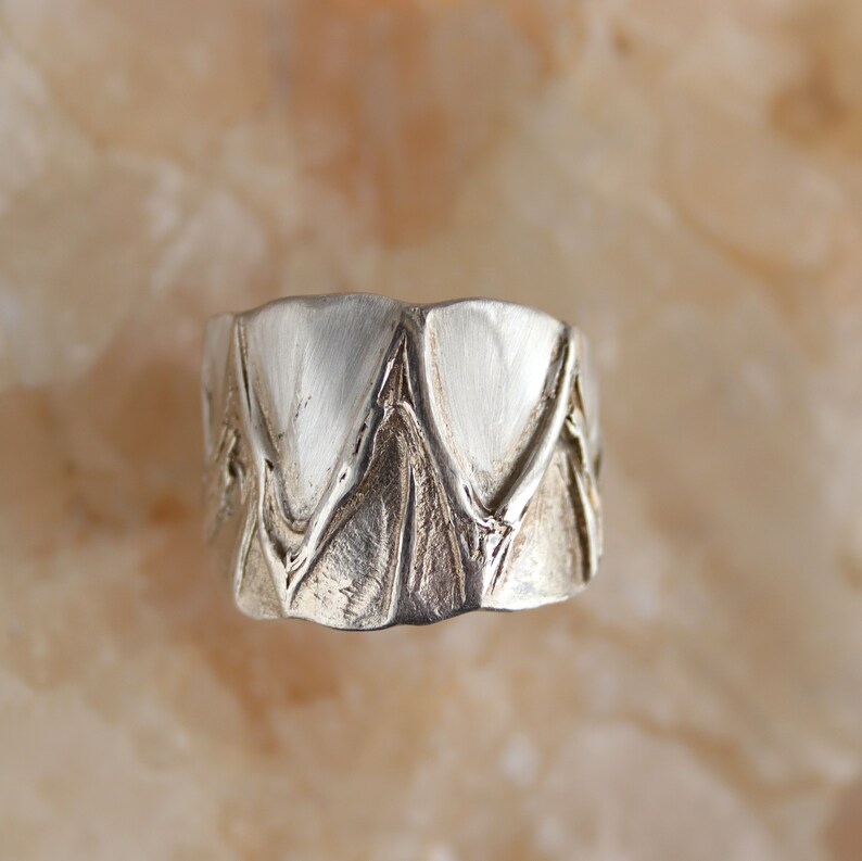 May include: A silver ring with a textured design featuring a series of overlapping triangles.
